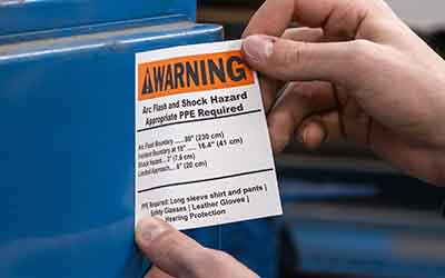 Operatore che applica un’etichetta di pericolo di folgorazione arancione su un macchinario.